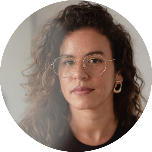 Foto de Maíra, que está de óculos, cabelos soltos de lado e olhando para a câmera com um leve sorriso.