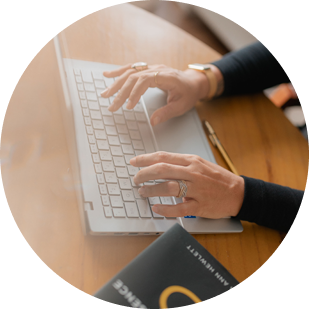 Foto das mãos de Maíra, digitando em um notebook branco e ao lado um livro preto que não dá para identificar o título.